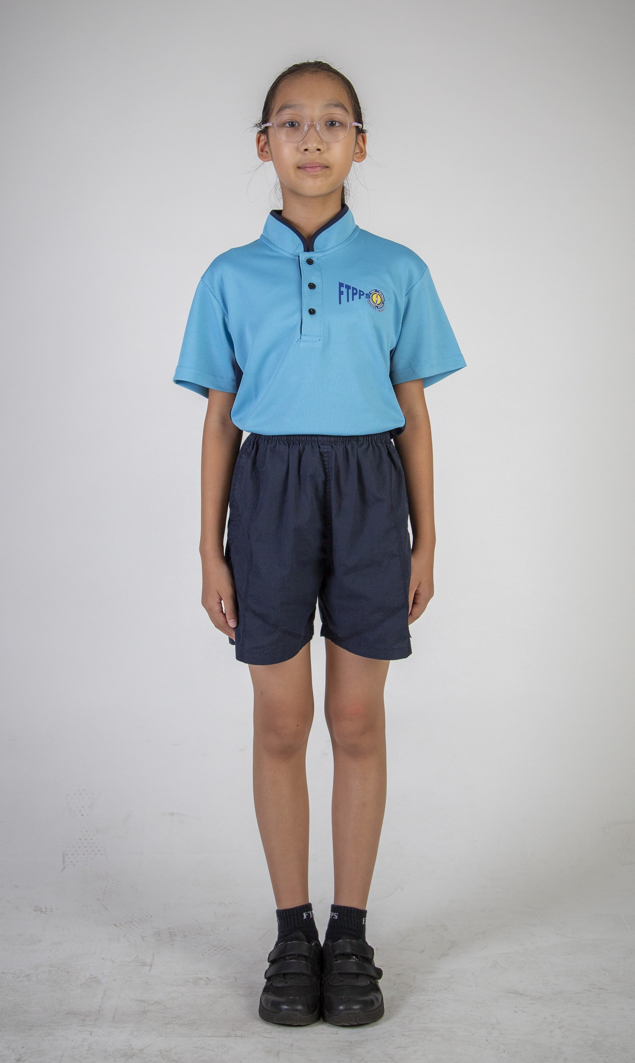 Front view of a student with tied-back hair and glasses, wearing a light blue sports polo shirt, navy shorts, black socks and black shoes.